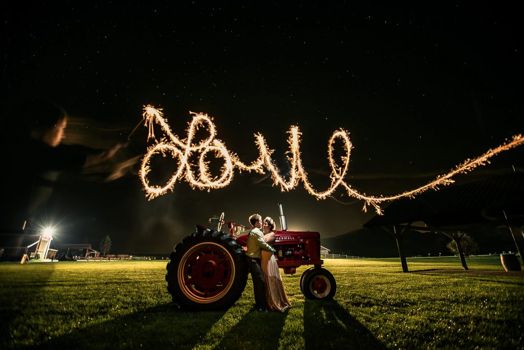 Fireworks spelling out "Love" above a couple standing together in front of a tractor at their Vermont farm wedding.