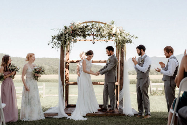Bride, groom, and wedding party standing under a rustic outdoor wedding arch