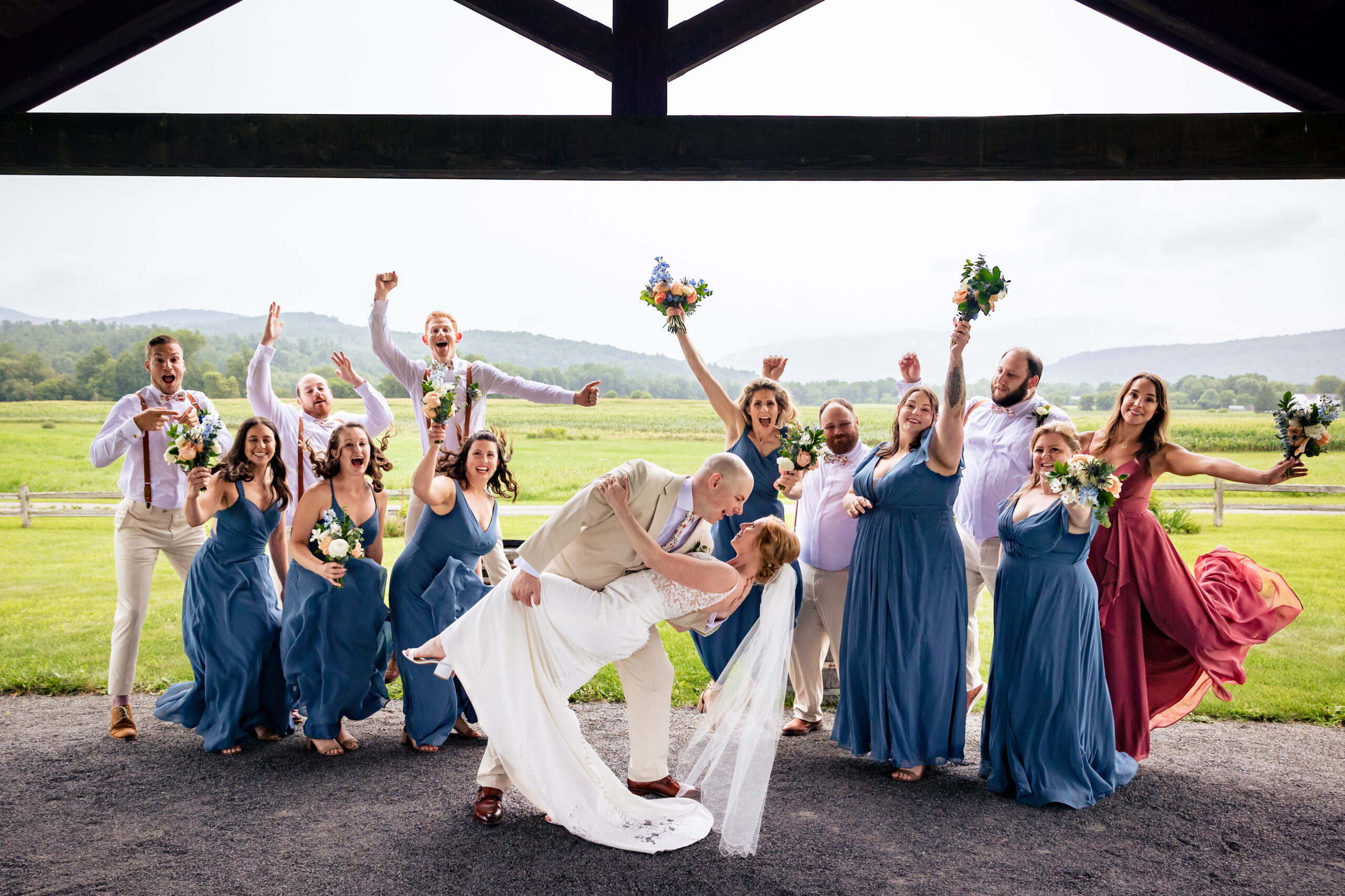 Husband dipping his wife at their summer wedding Venue in Vermont while the bridesmaids celebrate behind them in bright blue dresses.