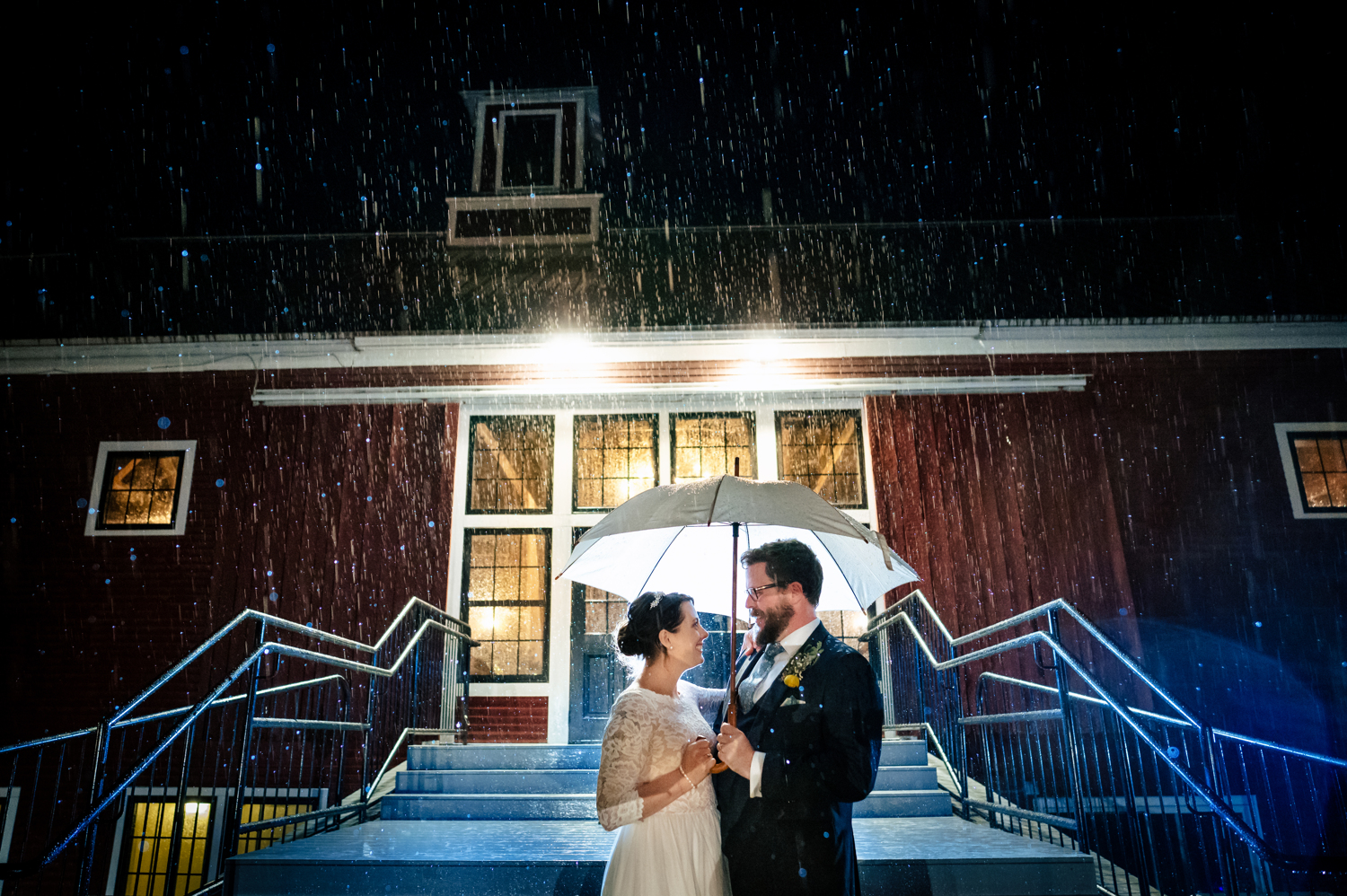 Anne and Jed standing and holding an umbrella in the rain in front of their wedding barn.