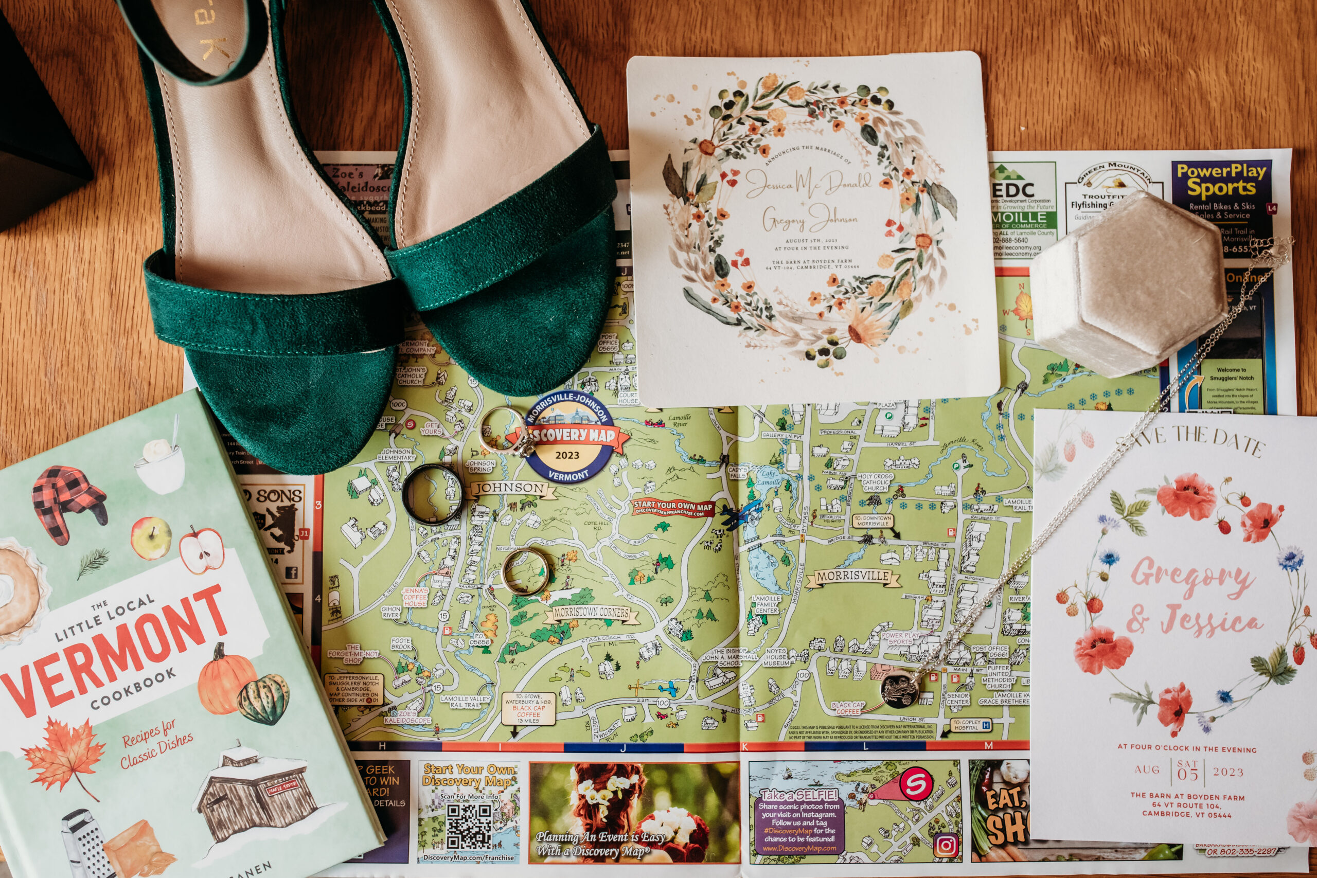 A pair of dark green suede heels, a save the date, a wedding invitation, a local map of northern Vermont, a Vermont cookbook, and wedding jewelry in a flat lay on a wooden table
