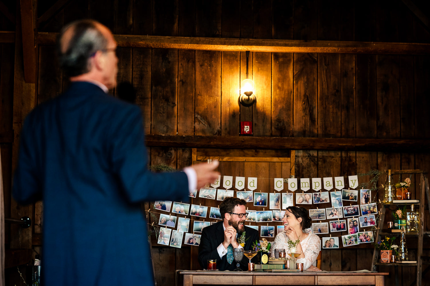 Anne and Jed sitting together at their table in their wedding barn while a man gives a speech.