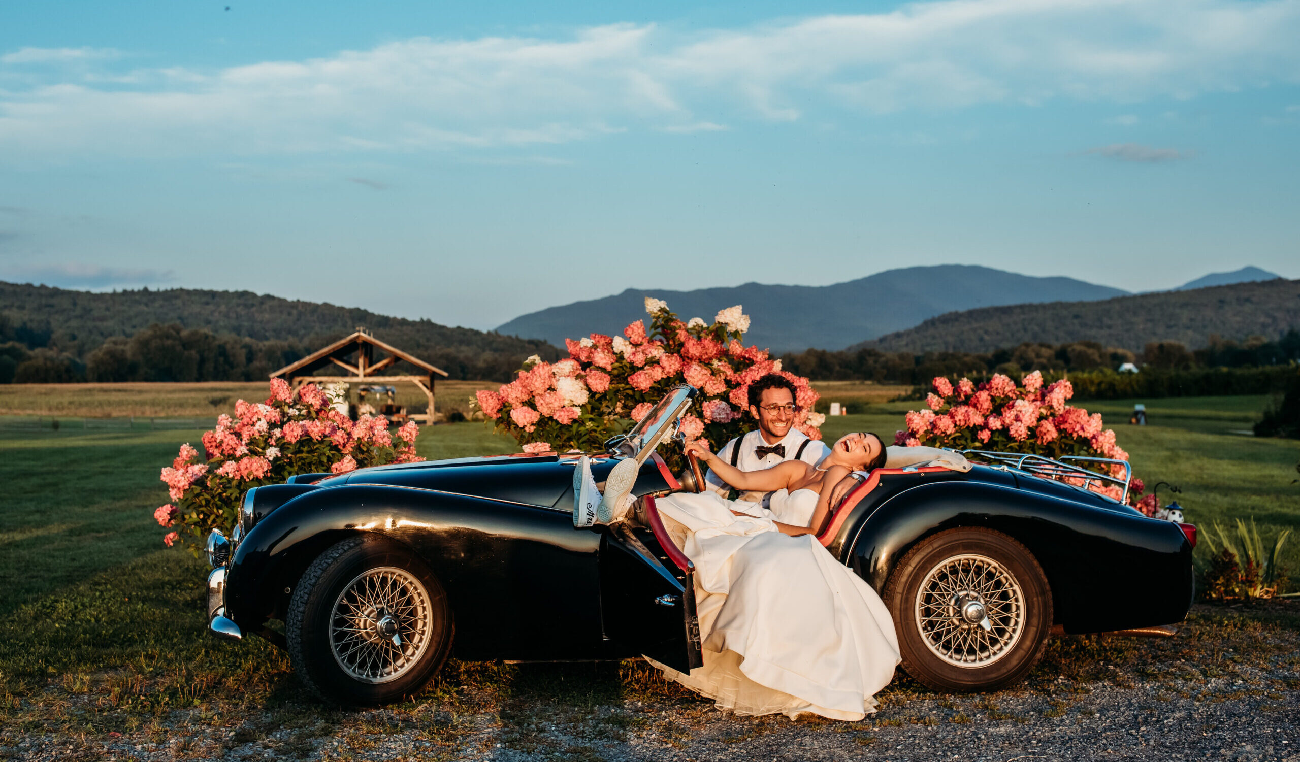 The bride and groom laughing and sitting together in a vintage car with mountain views behind them at their rustic wedding venue in Vermont