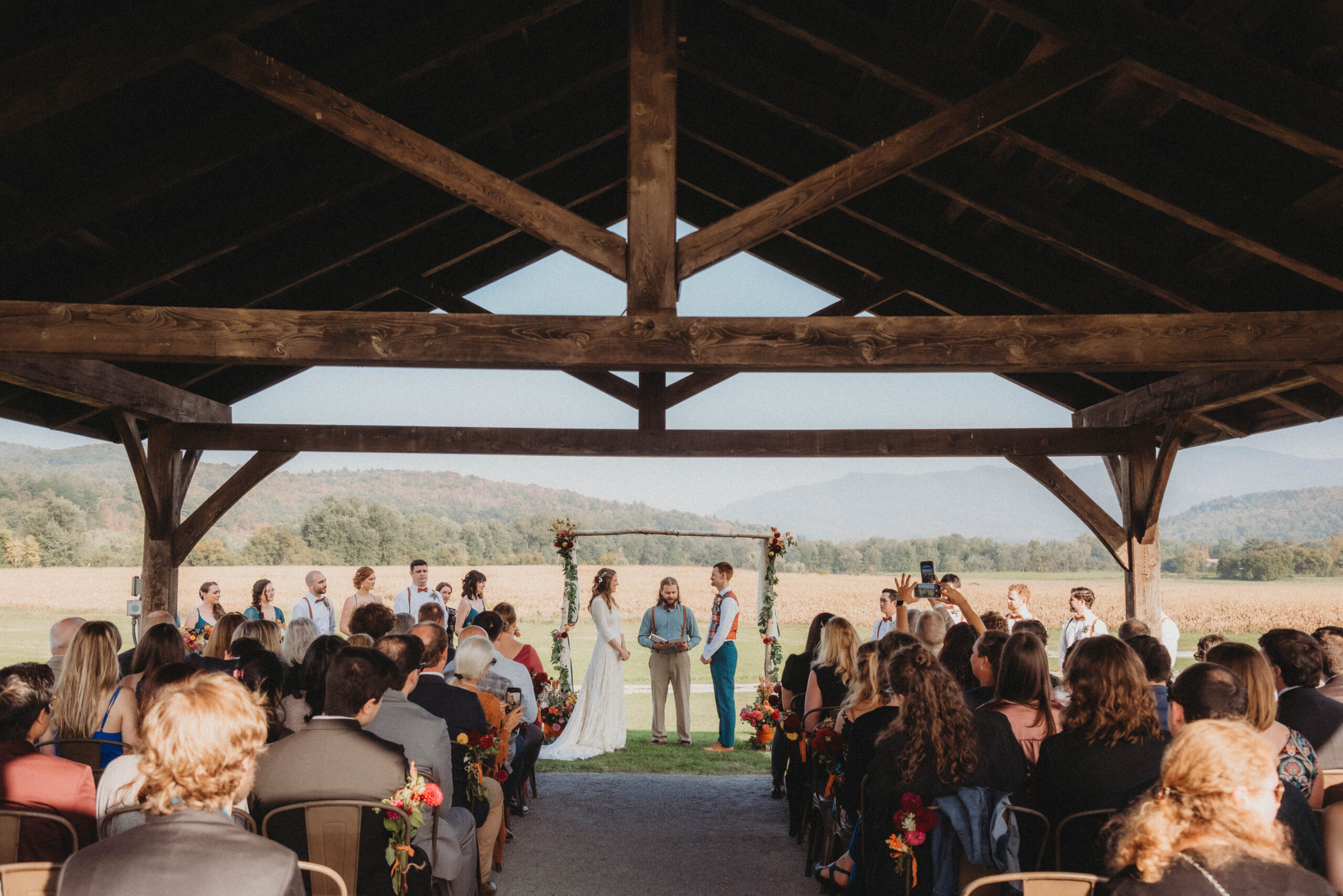 Emily and Sean standing together at their wedding arbor underneath the post and beam pavilion. Vermont mountains and a cornfield are in the background.