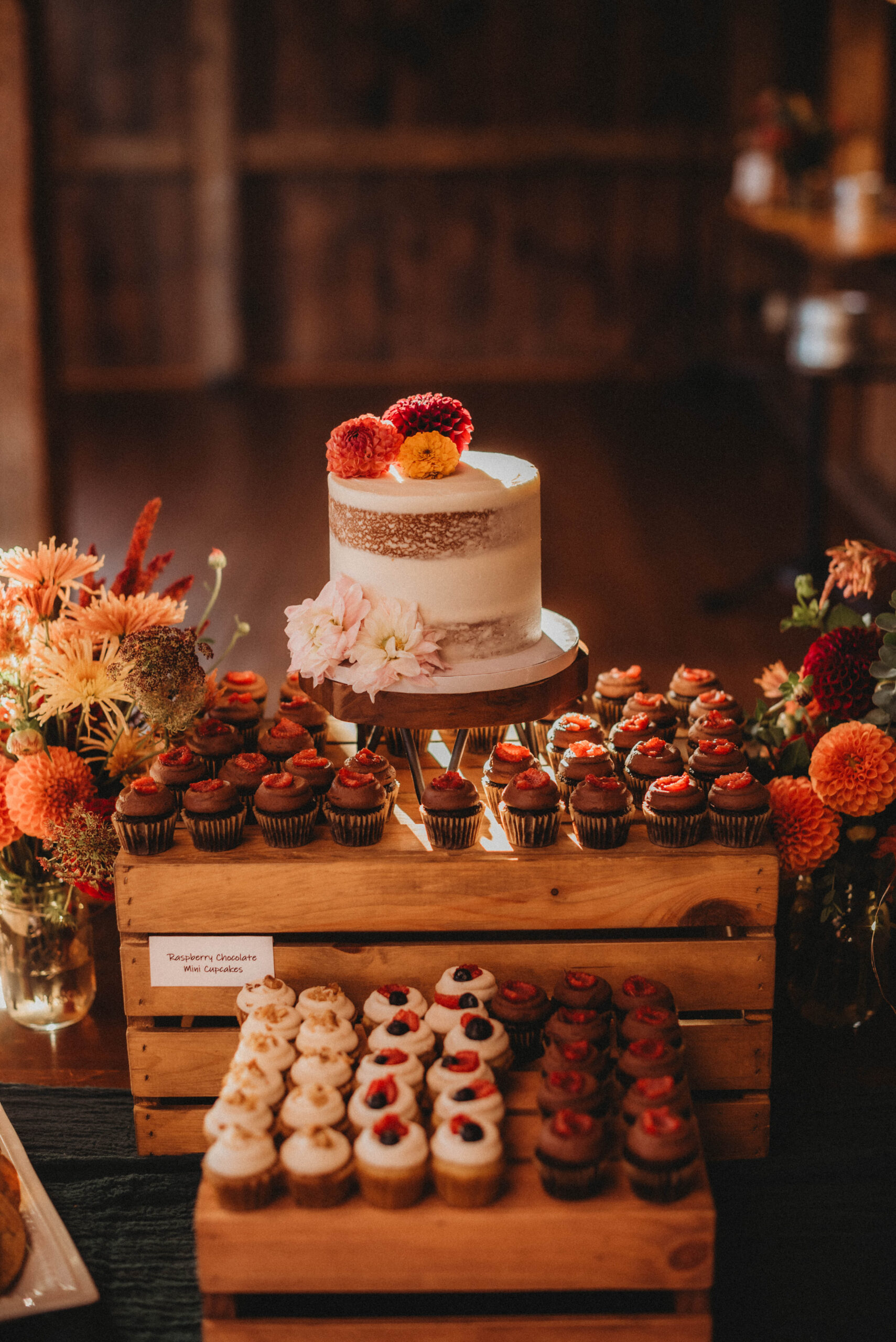 A small, rustic wedding cake sitting as the focal point of a dessert spread of cupcakes, cookies, and fall flowers