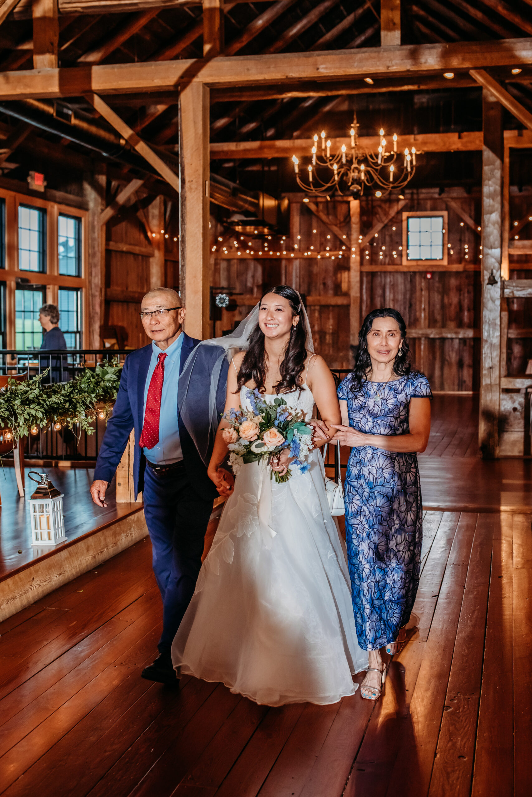 A bride being walked down the aisle of The Barn at Boyden Farm's rustic wedding barn by her two parents